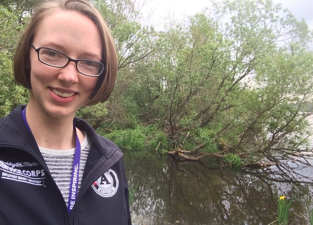 A woman smiles at the camera. There is water and a beaver lodge in the background.