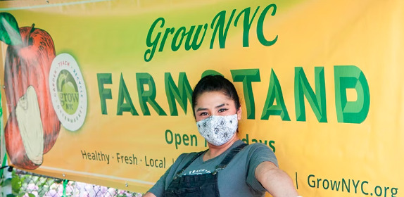 A GrowNYC Farm Stand worker smiling with a max on, carrying something off camera. They are in front of a large yellow banner that has "Grow NYC Farm Stand" written on it.