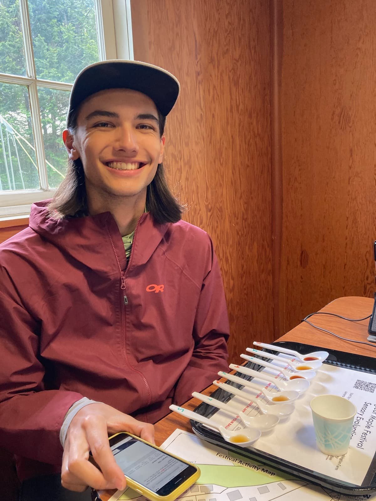Daniel wearing a hat smiling at a table. He has a tray with tasting spoons of sauces in front of him while his hand is holding his phone.