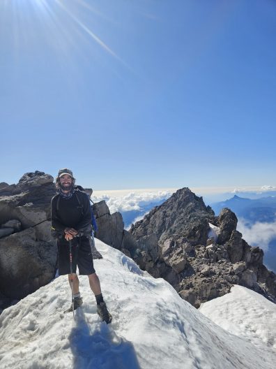 A person, smiling, stands on top a snowy peak.