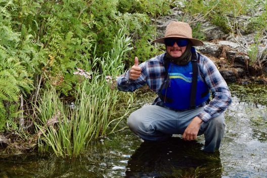 A person squats in a river near the bank, smiling.