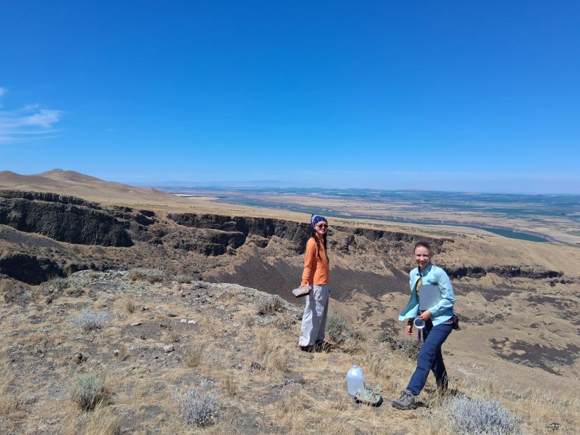 Two people standing on a plateau above a cliff, watering plants. 
